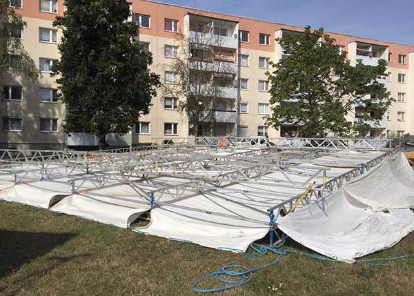Zeltaufbau mit Metallgerüst und Plane auf Wiese vor Wohnhaus bei sonnigem Wetter.