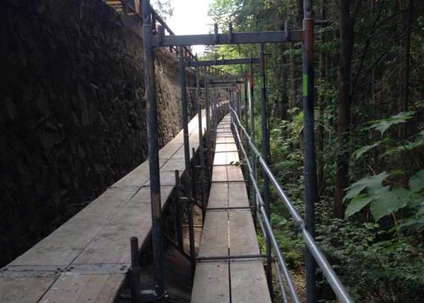 Gerüstbau an einer Steinmauer im Wald mit Holzbohlen und Metallstreben für Bauarbeiten.